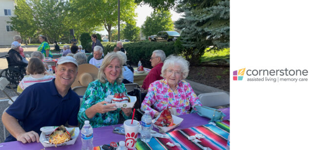 A group of three people are smiling and enjoying their meals at an outdoor table during the "Night to Unite" event at Cornerstone Assisted Living. The table is decorated with a colorful cloth, and other attendees are visible in the background, creating a warm and lively atmosphere.