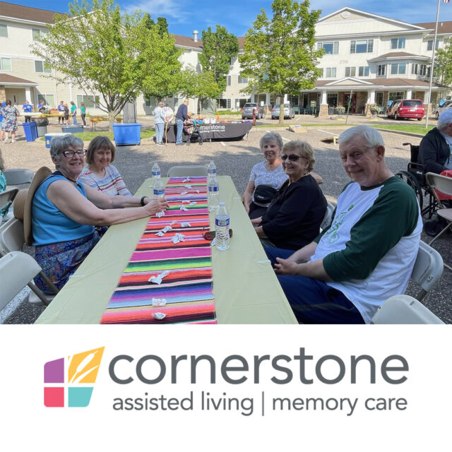 A group of people sitting around a long outdoor table, smiling and enjoying the beautiful weather during the "Night to Unite" event at Cornerstone Assisted Living. The colorful tablecloth adds vibrancy to the scene, with the Cornerstone logo visible in the background.