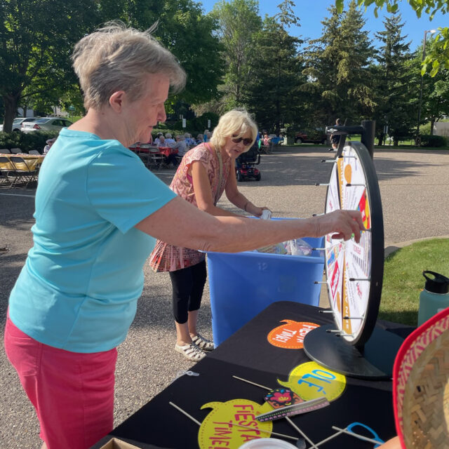 Two women interacting with a spinning prize wheel at the "Night to Unite" event. The event is taking place outside on a sunny day, with other tables and guests visible in the background.